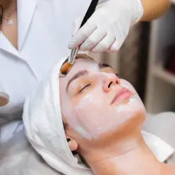 Mulher jovem aplicando creme no rosto durante sessão de limpeza de pele para promover hidratação e rejuvenescimento em uma clínica de estética